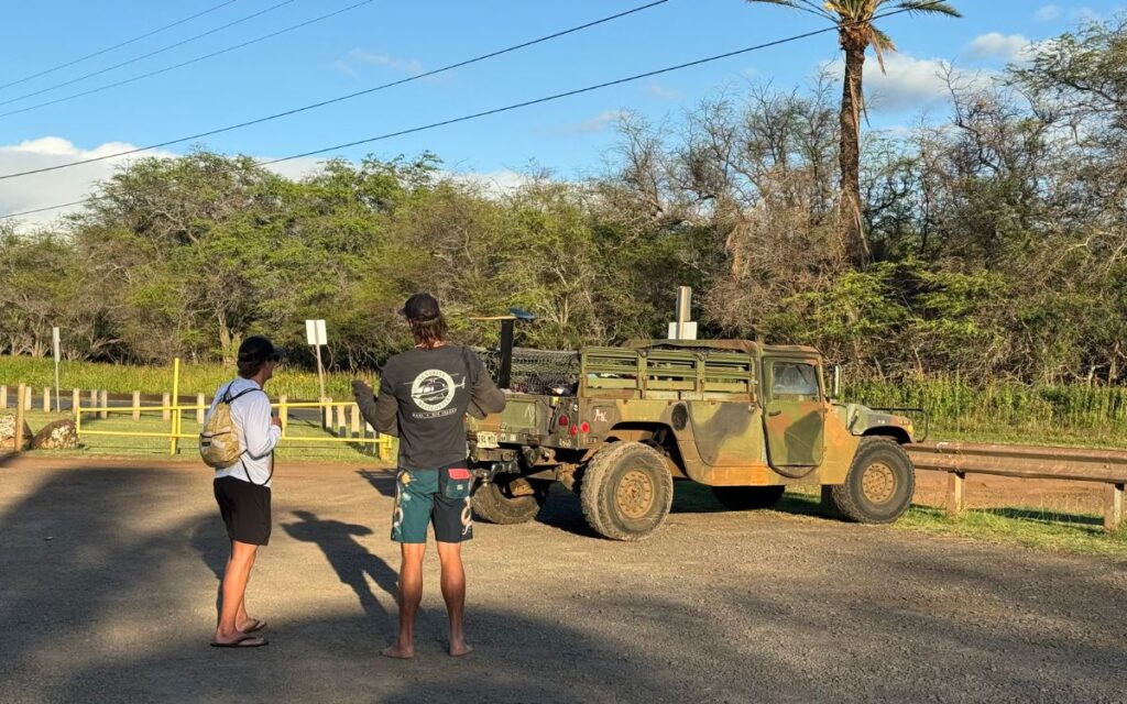 Two individuals from behind observing an old military jeep parked on a dirt road, surrounded by rustic scenery and tropical foliage.