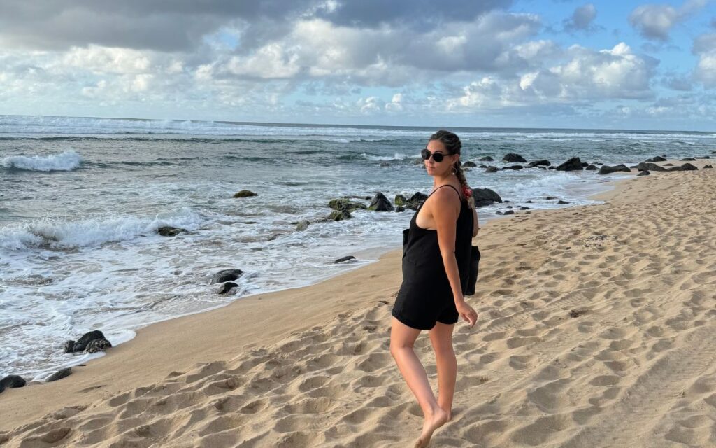 A woman in a black dress walks along a sandy Hawaiian beach, with waves and rocks in the background under a cloudy sky