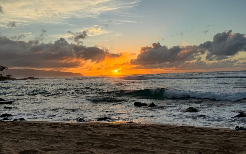 Glorious sunset with vibrant hues of orange and purple over a churning sea, viewed from a sandy shore with rocks.