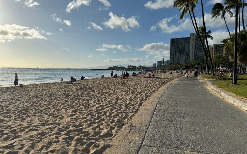A sandy beach path with visitors enjoying a sunny day, clear blue waters, and a city skyline in the distance.