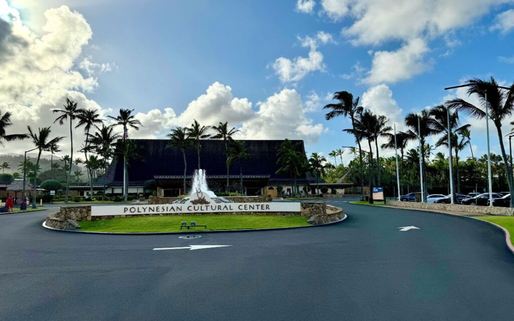 Polynesian Cultural Center entrance with its name on a sign, a large traditional structure behind, surrounded by palm trees under a partly cloudy sky.