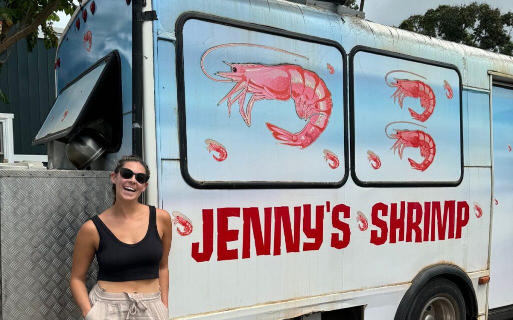 Side of 'Jenny's Shrimp' food truck with illustrations of red shrimp, with a delighted woman standing beside it, under a clear blue sky