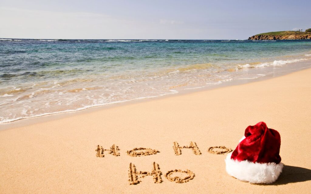 A beach scene during the holiday season featuring "Ho Ho Ho" written in the sand with a Santa hat nearby, the gentle surf in the background, under a clear blue sky.