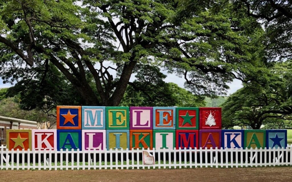 Brightly colored blocks spelling out "MELE KALIKIMAKA" are displayed in front of a large tree. The blocks are arranged in a festive pattern, with stars and holiday symbols, all behind a white picket fence.