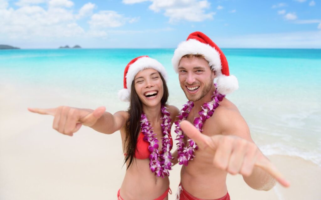 A joyful couple wearing Santa hats and leis, posing with a thumbs up on a tropical beach, smiling broadly and pointing at the camera, under a clear sky with a serene ocean backdrop.