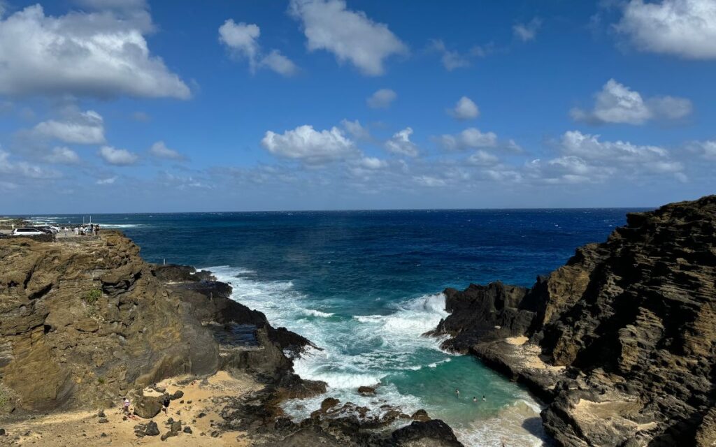 A coastal scene featuring rugged, rocky cliffs surrounding a small cove with clear blue waves crashing against the shore. In the background, the expansive ocean meets a partly cloudy sky, while a few people can be seen exploring the rocks and swimming in the water.